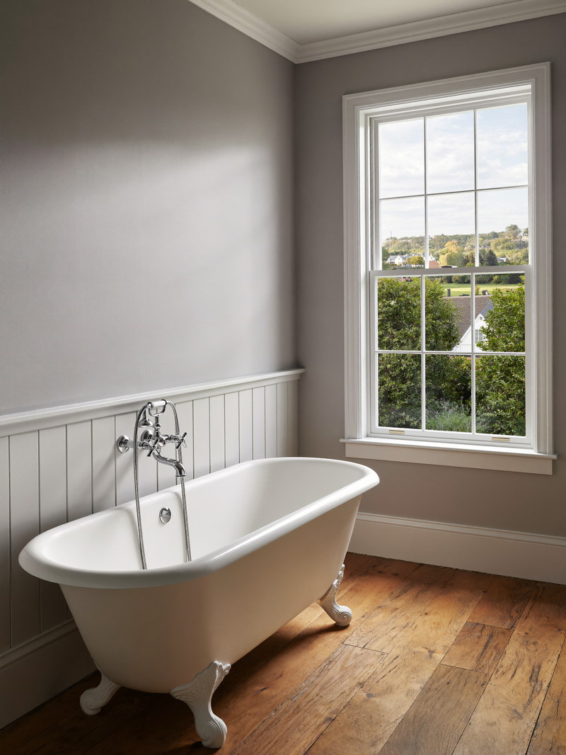 A renovated colonial-home bathroom with soft gray walls, white clawfoot-style tub with plain smooth feet, white wainscot, wide-plank wood floor