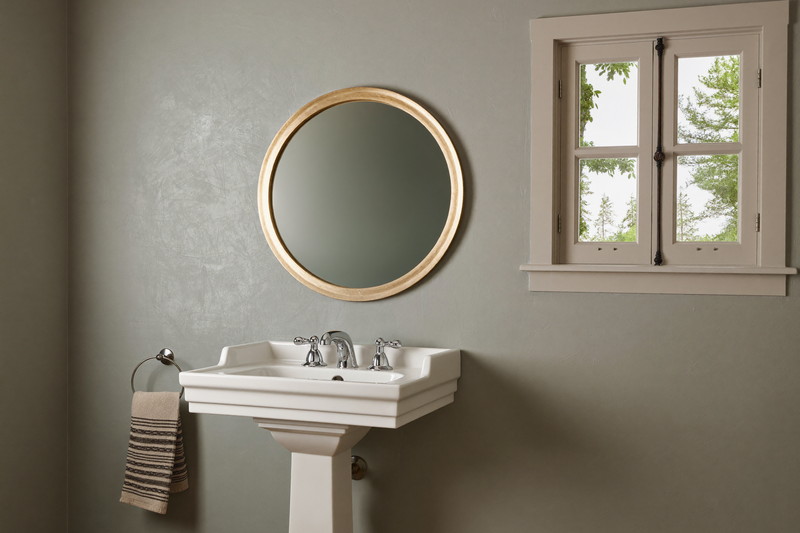 A compact powder room in a New England colonial: soft sage-gray walls, white pedestal-style washstand, round nickel-framed mirror