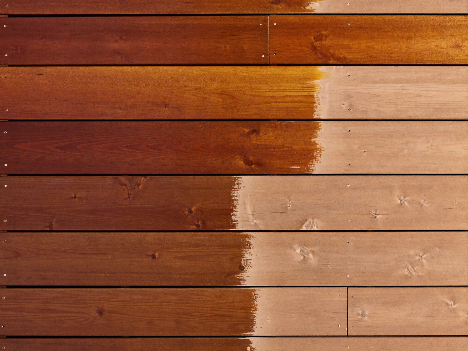 Overhead detail photograph of a deck mid-restaining: rich warm stain covering half the cedar boards meeting bare sanded wood at a crisp brushed line along on