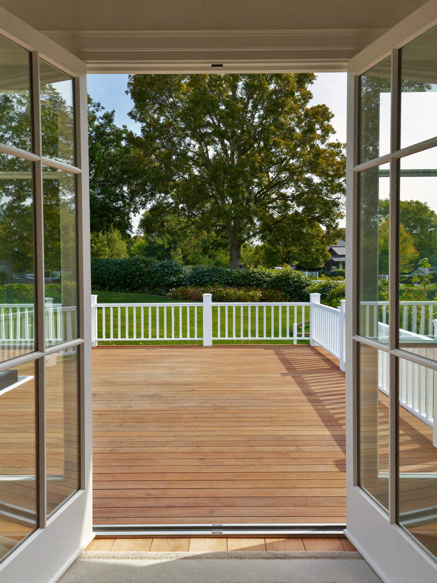View through open french doors from a colonial family room onto a warm cedar deck with white railing and a leafy suburban back yard beyond