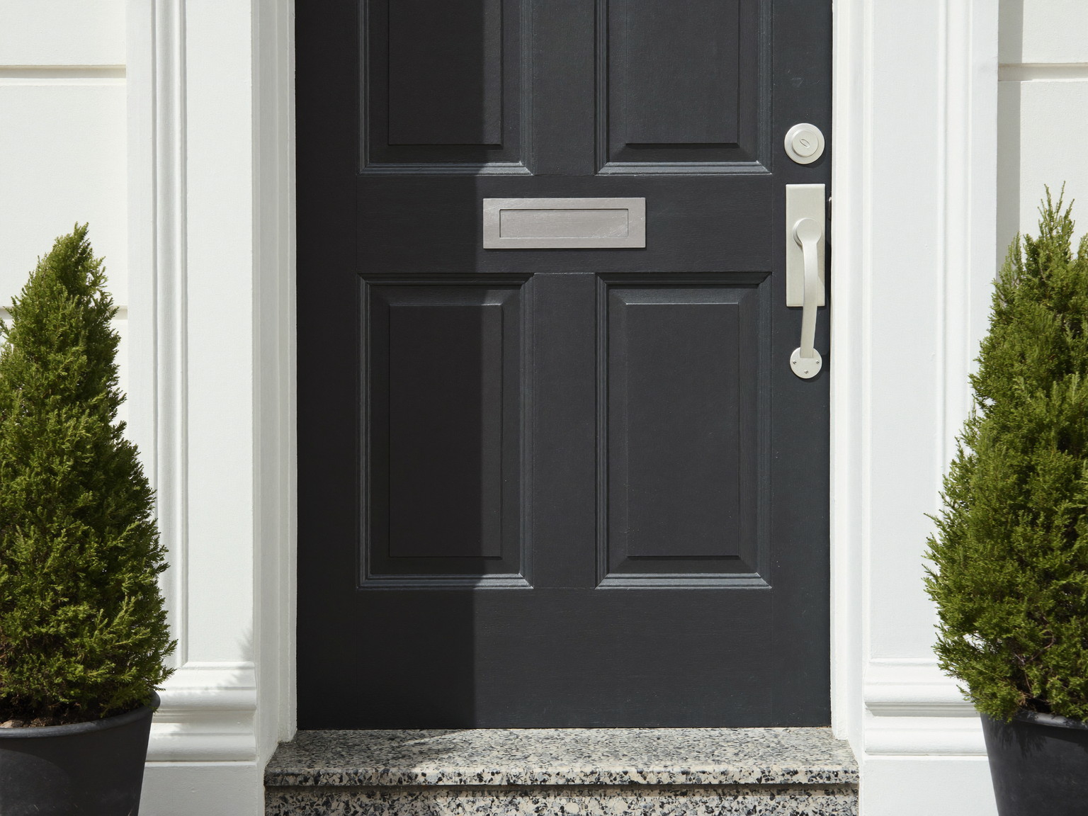 A cared-for colonial entry: repainted paneled door in deep charcoal, repaired white casing, polished plain nickel hardware, swept granite step, potted boxwood