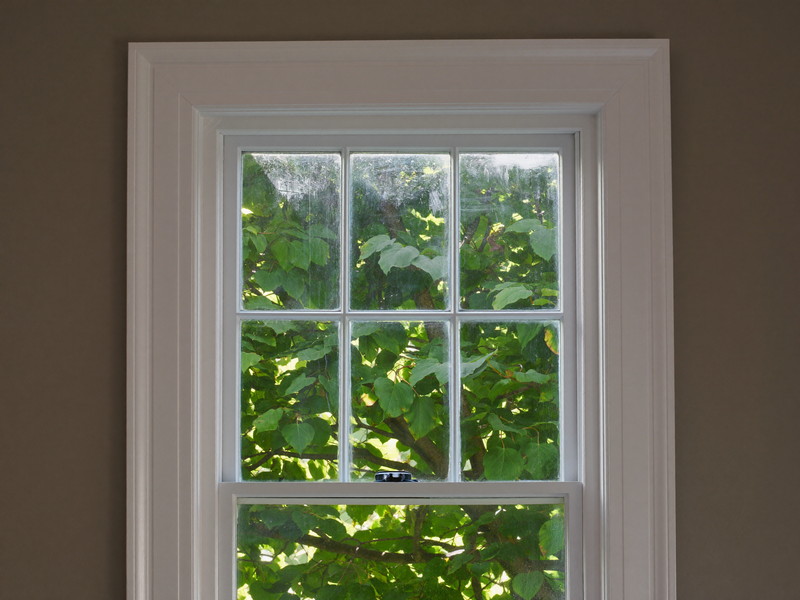 A restored double-hung window sash in a colonial home: fresh white paint, clean glazing, original wavy glass catching light, view of green leaves outside