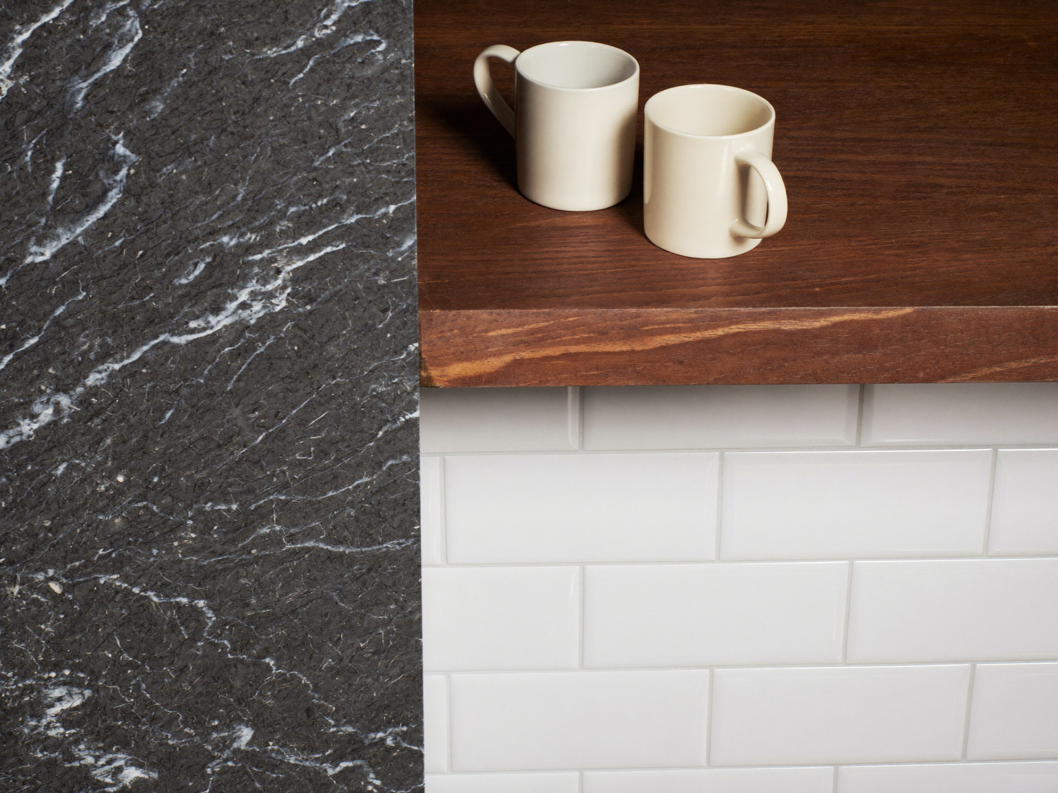 Macro photograph where a dark honed soapstone counter meets a glossy white subway tile backsplash with a slim oak shelf above holding plain ceramics