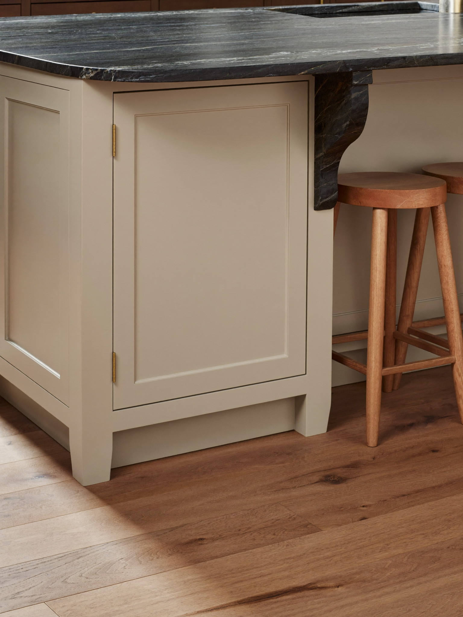 Vertical detail photograph of a cream shaker kitchen island end with applied panel, dark soapstone overhang, plain wooden stools, wide-plank oak floor