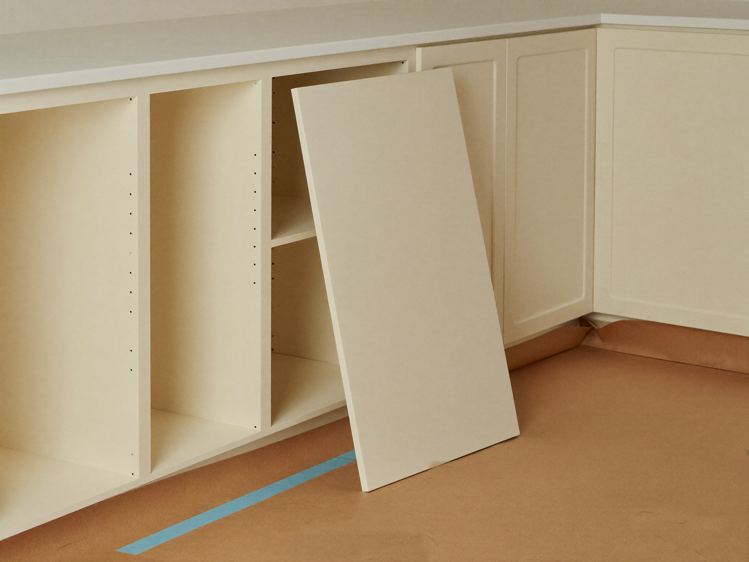 A suburban kitchen renovation in progress: cream inset cabinets installed and shimmed level, unmarked doors leaning wrapped nearby