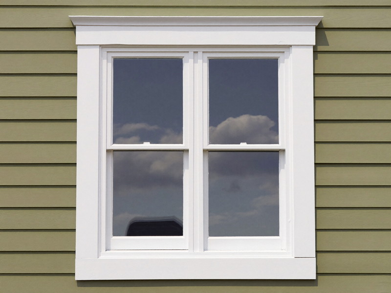 A freshly repainted double-hung window and casing in bright white against muted sage clapboard siding, crisp caulk lines, soft sky reflection in the glass