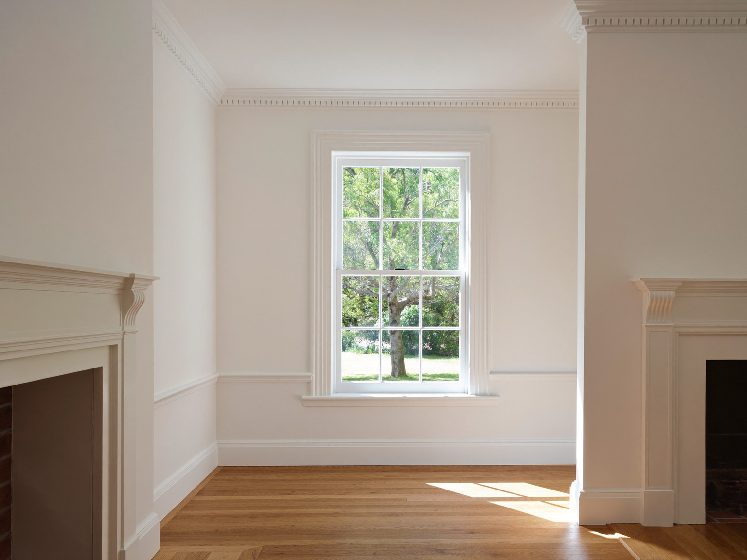 A finished colonial living room with fresh warm white walls, crisply painted crown molding and window trim, oak floor glowing in afternoon light, simple mantel