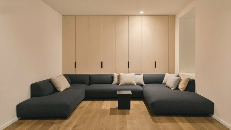 The finished lower level of a comfortable Medway Massachusetts colonial, softly lit: warm white walls, pale wood built-in media wall with plain closed cabinets, a deep charcoal-navy sectional with light gray cushions, a small completely bare dark side table, wide plank flooring