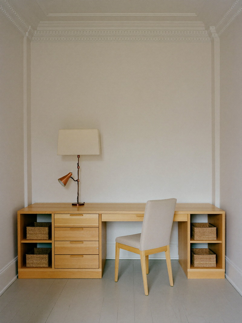 A comfortable lower-level home office finished in a renovated colonial basement: built-in pale wood desk with plain drawer faces against a smooth bare warm white wall, simple upholstered chair, copper task lamp with a plain shade, each open shelf holding a single plain woven bin