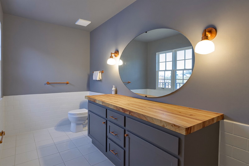 A calm finished suburban bathroom in soft evening light: brushed copper sconces glowing beside an oval mirror, blue-gray vanity with a completely bare