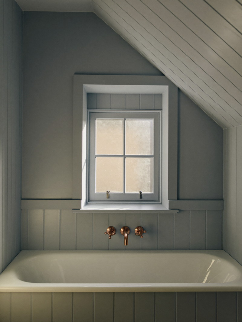 A renovated bathroom under the sloped ceiling of a Medway cape: white tub, soft blue-gray paneling, brushed copper fixtures, small window with frosted