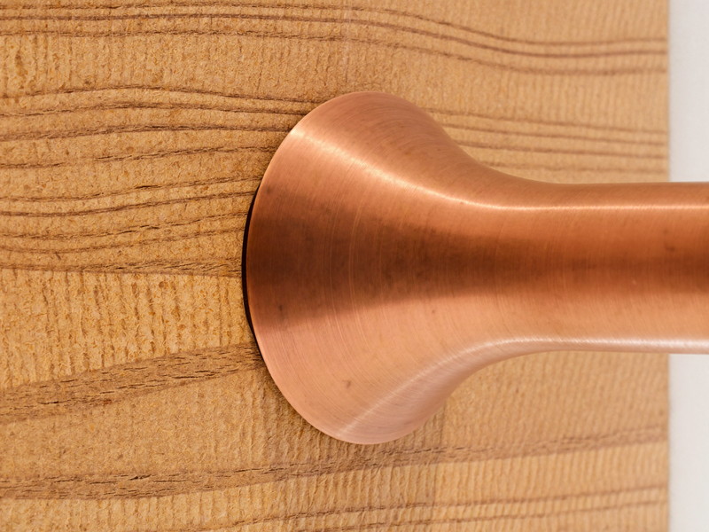 Macro photograph of warm butcher-block wood grain meeting the plain rounded base of a brushed copper faucet seen from the side, the metal completely bare unmarked with no engraving, shallow depth of field, bright natural light