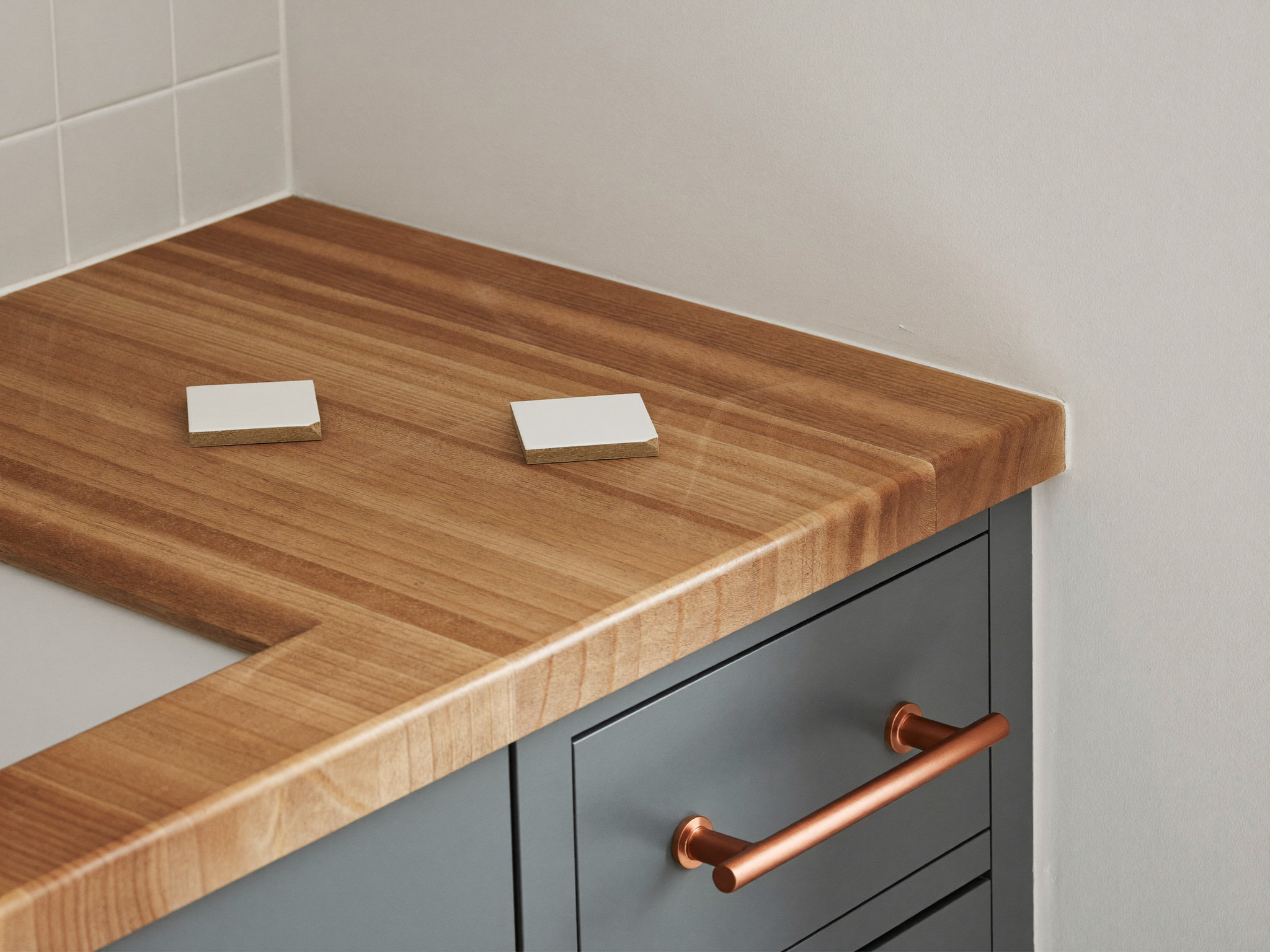 A corner of a blue-gray bathroom vanity in soft daylight: the warm butcher-block top meeting a warm white wall, two loose warm white square tiles resting on the bare wood surface, a brushed copper bar pull on the drawer below, every surface completely blank and bare