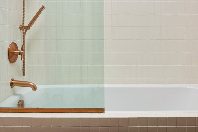 A tidy tub and shower combination in a suburban family bathroom: white tub, warm white square tile surround, brushed copper fixtures, clear glass screen, every ledge and edge completely bare, crisp and clean