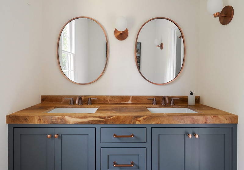 A double soft blue-gray vanity with butcher-block top under two oval copper-framed mirrors, brushed copper sconces, warm white walls, comfortable suburban