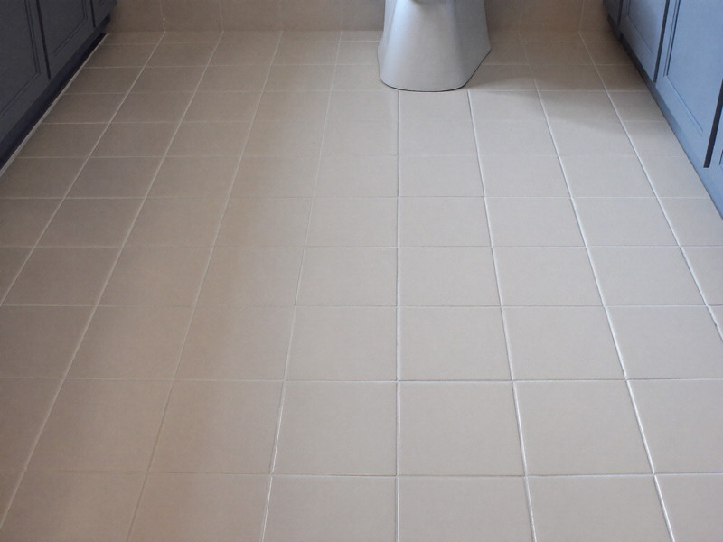 Large-format warm white floor tile in a renovated suburban bathroom, plain field tile only, every tile flat and even, foot of a blue-gray vanity visible,
