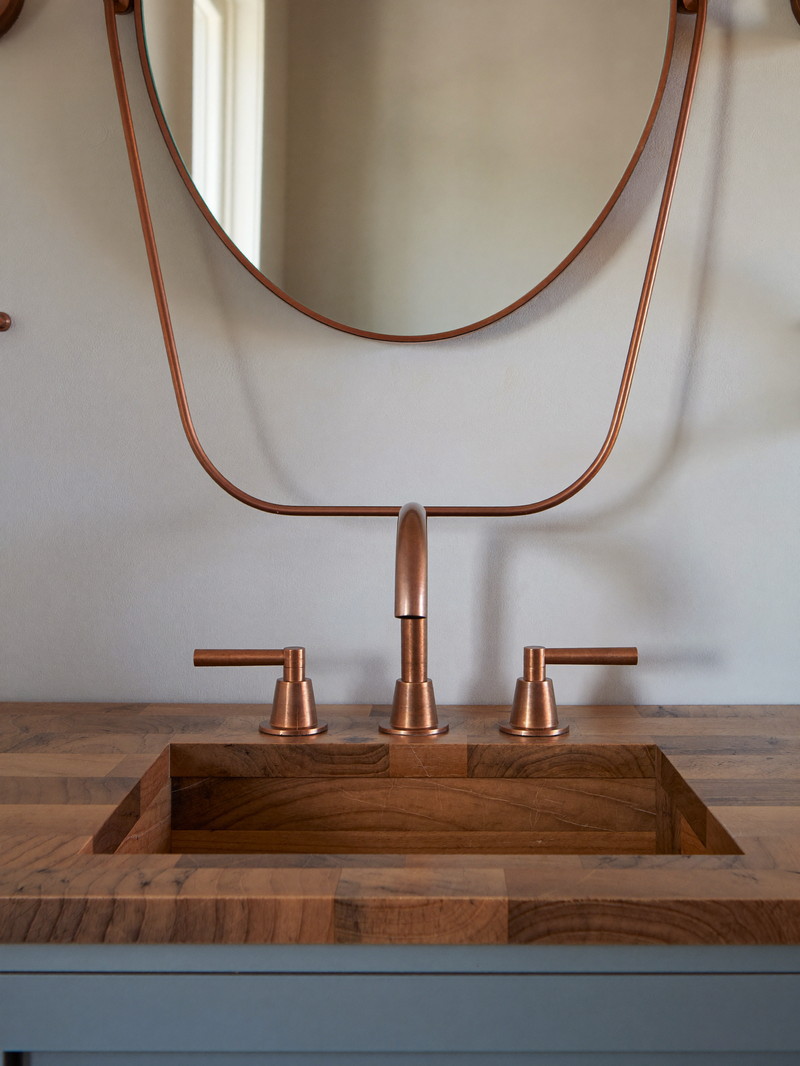 A soft blue-gray painted bathroom vanity with a warm butcher-block wood top and brushed copper faucet with smooth lever handles, oval copper-framed mirror