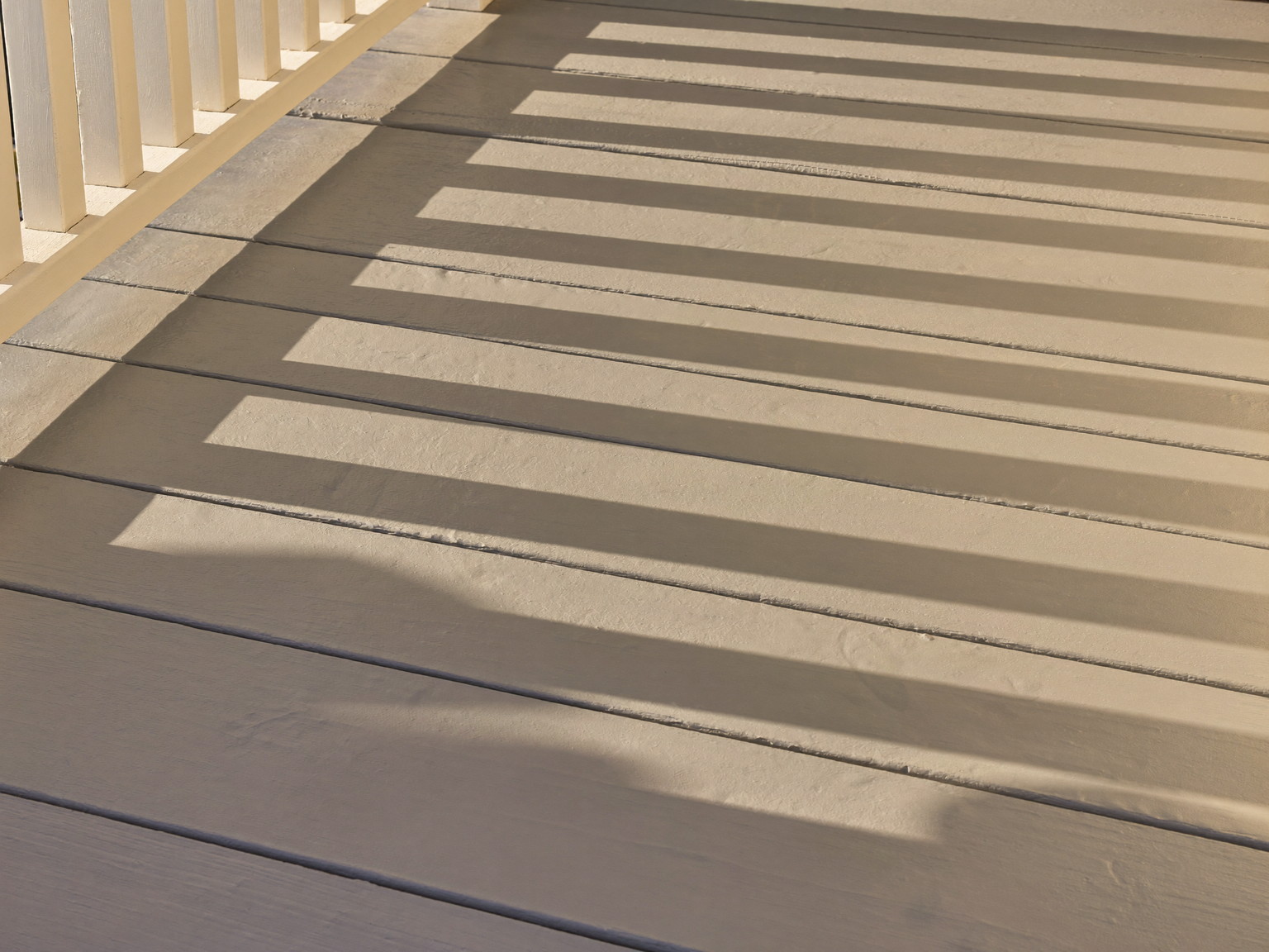 A freshly painted gray porch floor drying in warm afternoon light: smooth even surface with a soft sheen, crisp white baluster shadows falling across the