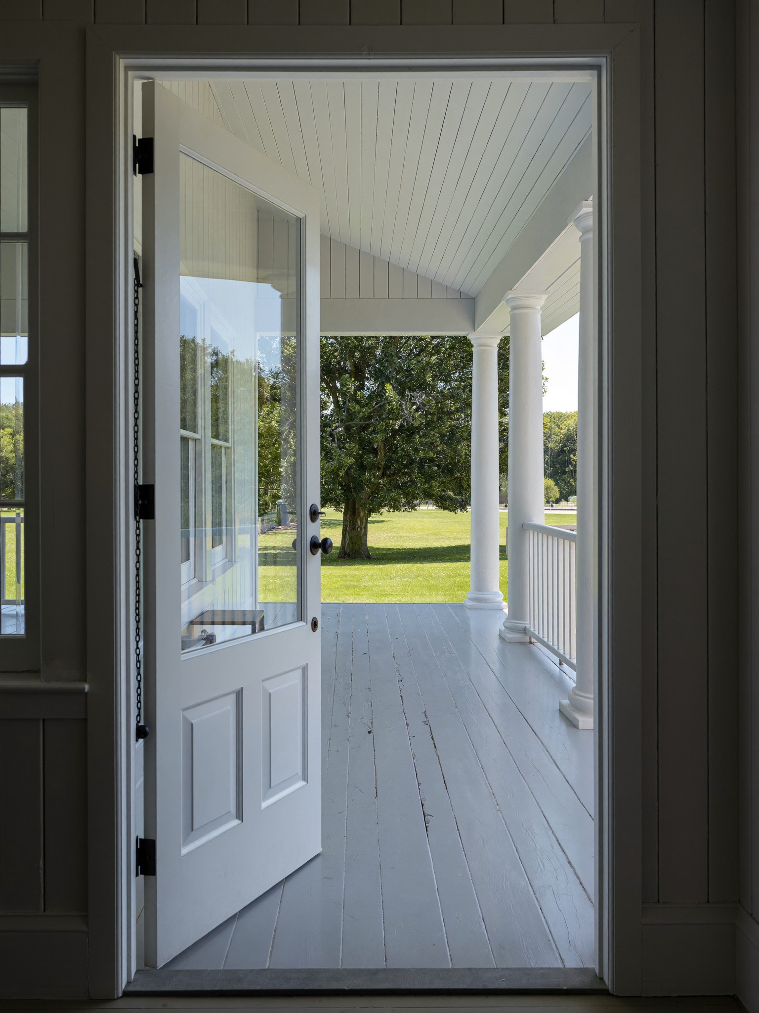 Tall photograph through an open white front door onto a restored farmer's porch: fresh gray painted floor, white columns and railing, green lawn and