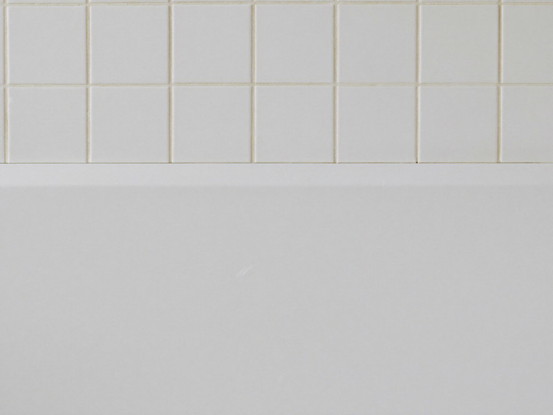 A clean bright white bathtub edge meeting warm white square wall tile, the joint freshly finished, smooth and tidy, completely bare tub edge, soft daylight