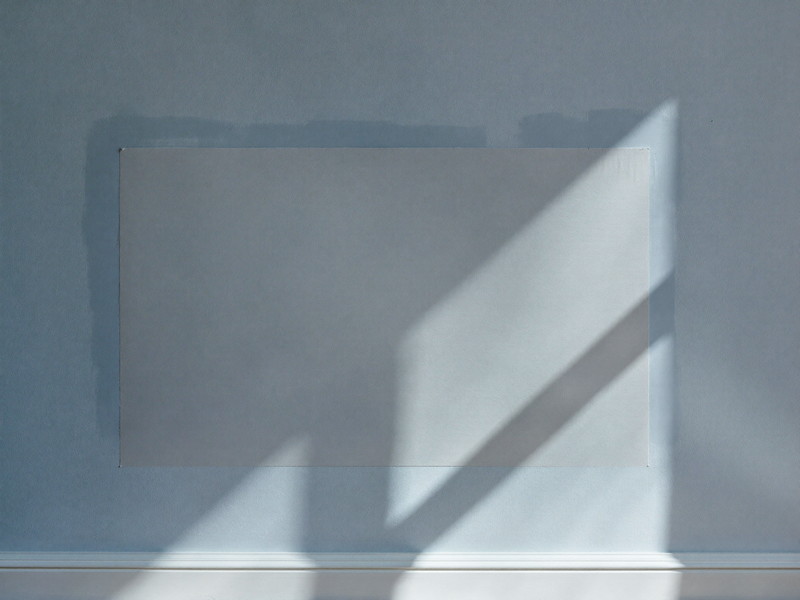 A freshly patched wall in a comfortable home: the repaired rectangle sanded perfectly flush and smooth, visible only as a faint matte patch on the soft blue-gray painted plaster wall under raking window light, crisp white baseboard below