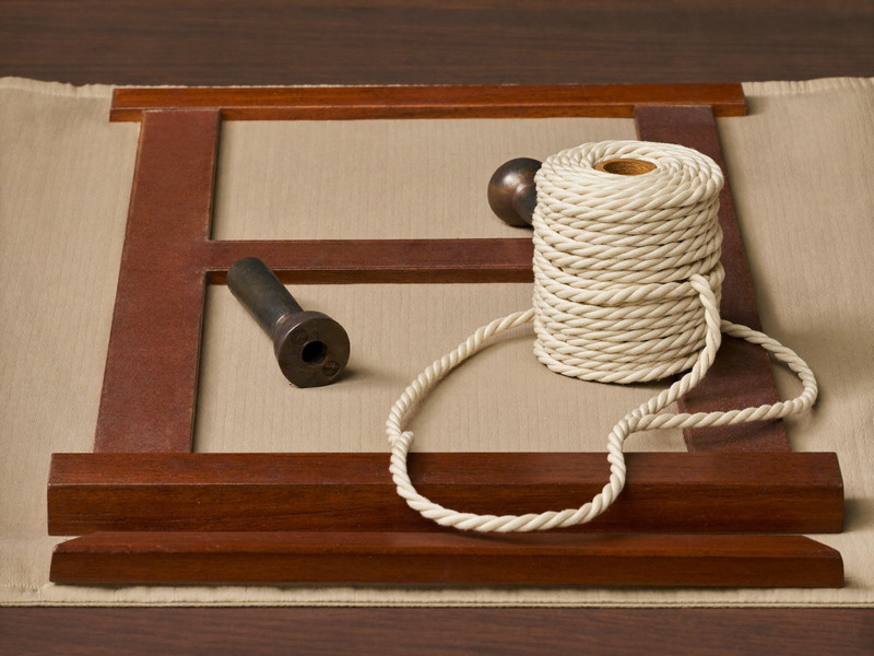 Resting on a padded workbench, a colonial double-hung wooden sash mid-service: soft twisted cotton sash cord in a loose coil beside plain smooth iron sash weights, no markings, soft workshop light