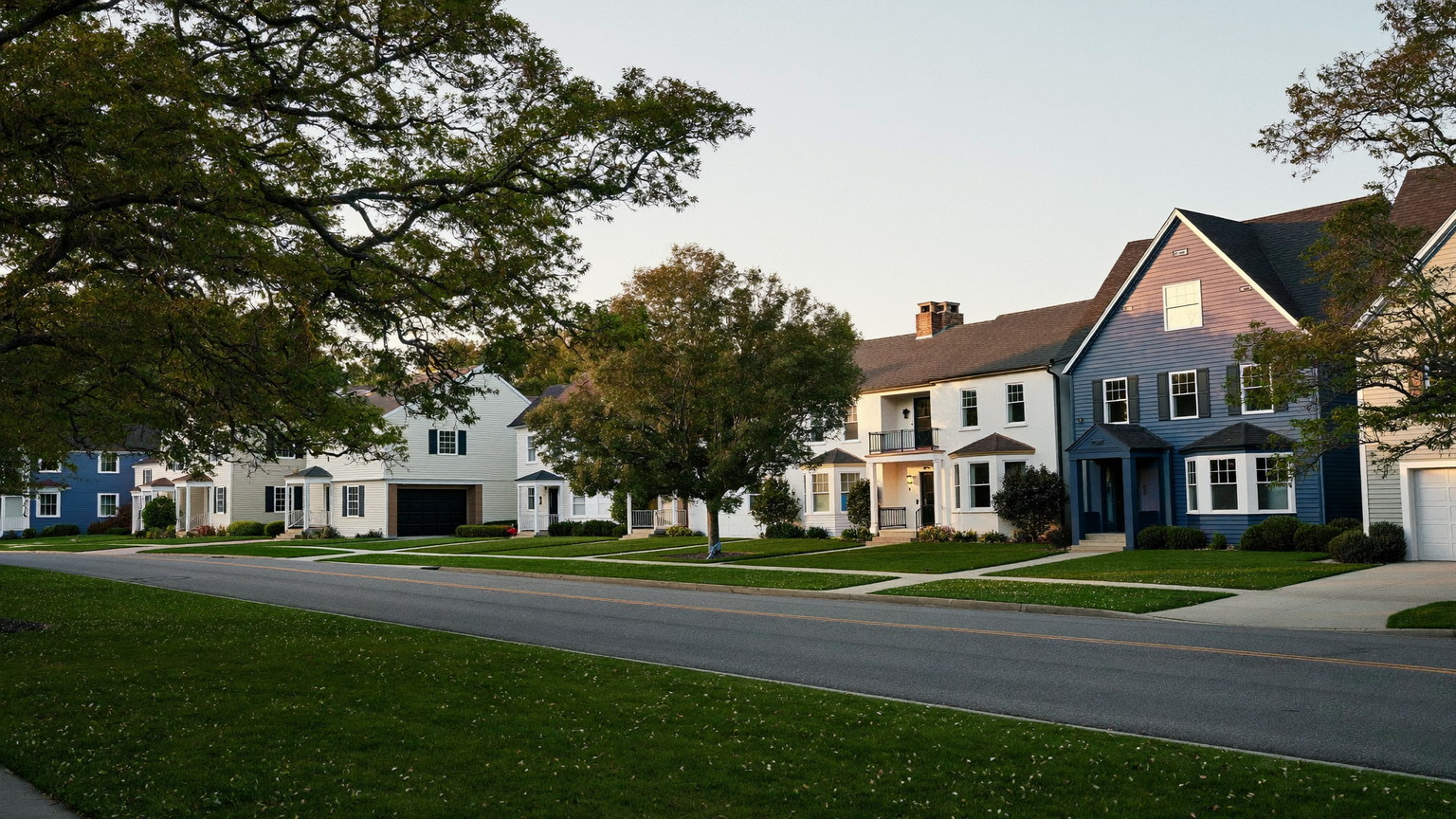 A quiet Medway Massachusetts residential street in early evening: comfortable established detached colonials, capes, and split-levels set apart on deep