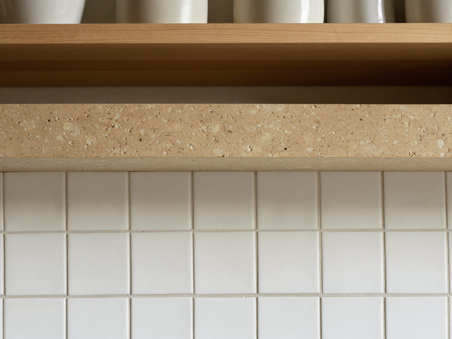 Macro photograph of a honed stone counter edge with soft fossil flecks meeting a warm white square tile backsplash, a slim pale wood shelf above holding plain white ceramics, soft window light