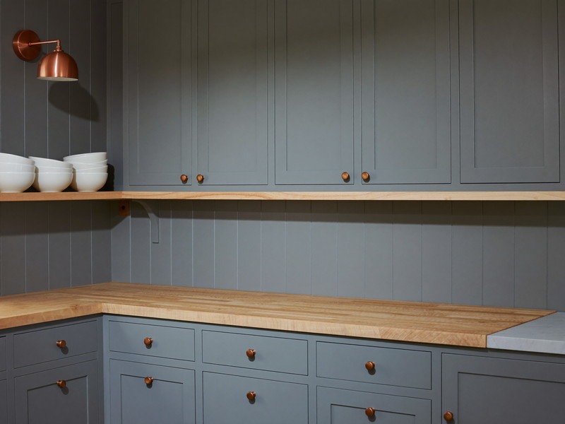 A finished suburban kitchen corner at evening: soft blue-gray shaker cabinets with brushed copper knobs, a completely bare butcher-block counter with nothing on it, pale wood shelf holding only plain white bowls, lit warm by a small copper dome sconce, calm and inviting