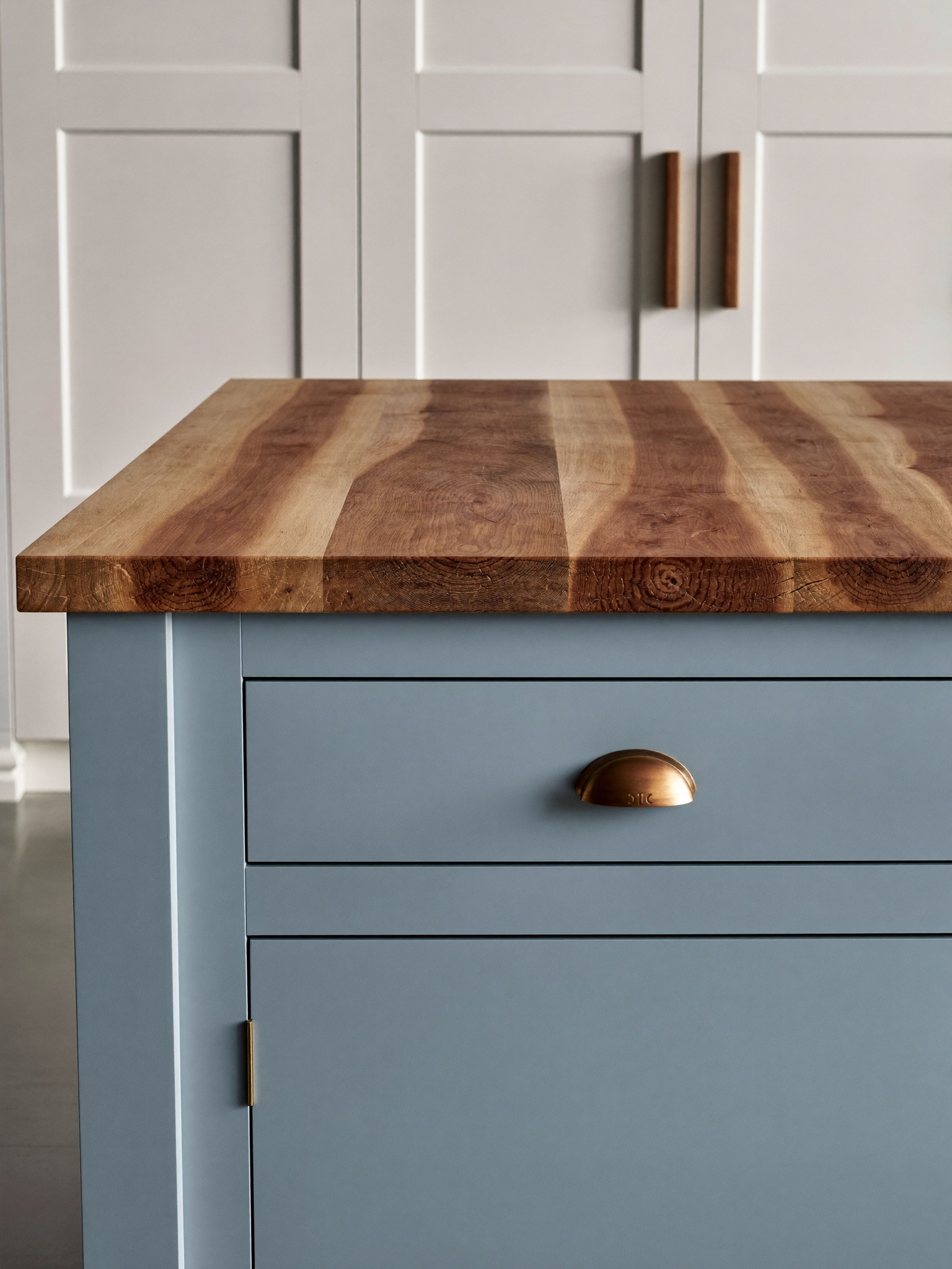 A soft blue-gray kitchen island with a thick warm butcher-block wood top with visible wood grain, brushed copper cup pulls with smooth faces, warm white shaker cabinets behind, natural light