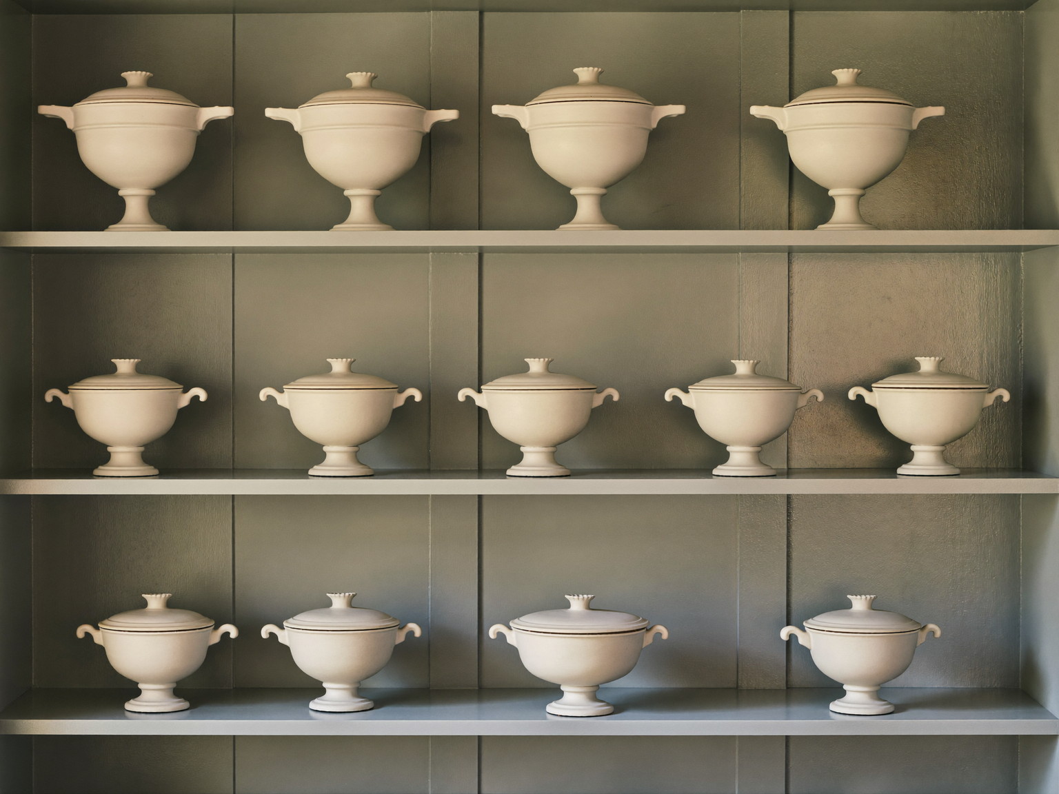 Inside a comfortable colonial's pantry: soft blue-gray painted shelves lined with plain white lidded ironstone tureens, every piece completely plain and bare, warm light