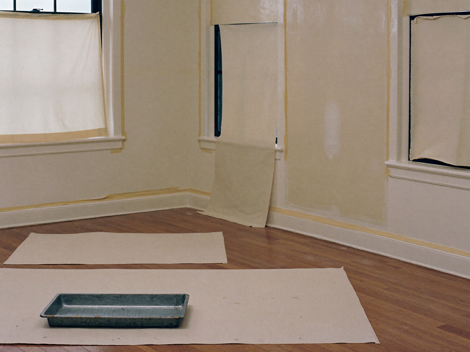 Interior painting prep, seen from across a comfortable colonial room: clean canvas drop cloths spread over the oak floor, window casings masked with plain