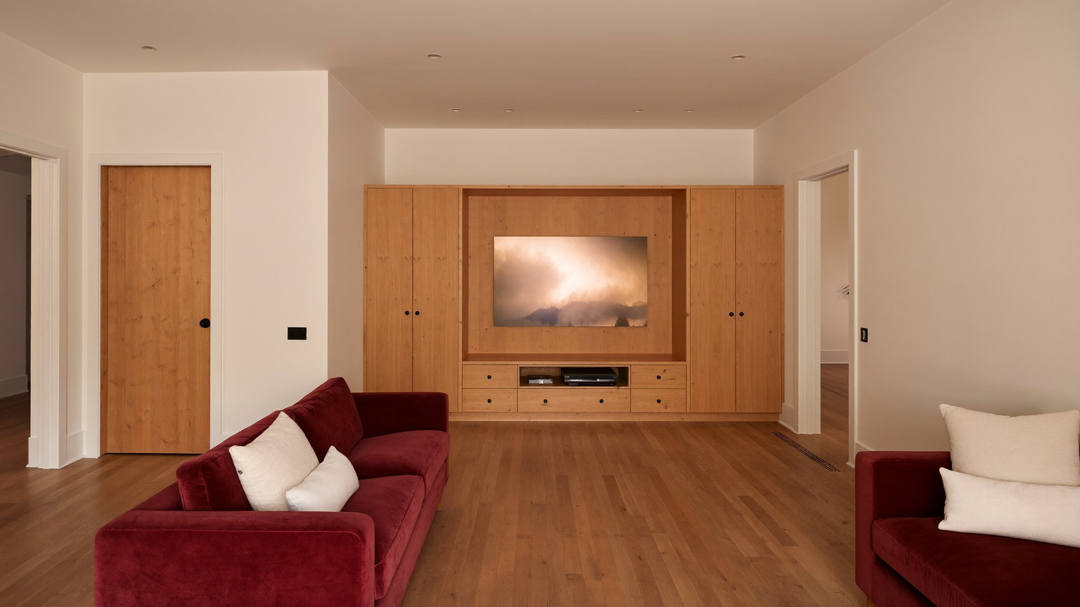 The finished lower level of a well-kept Millis Massachusetts colonial, warm and lamplit: soft cream walls, warm pine built-in media wall with plain closed