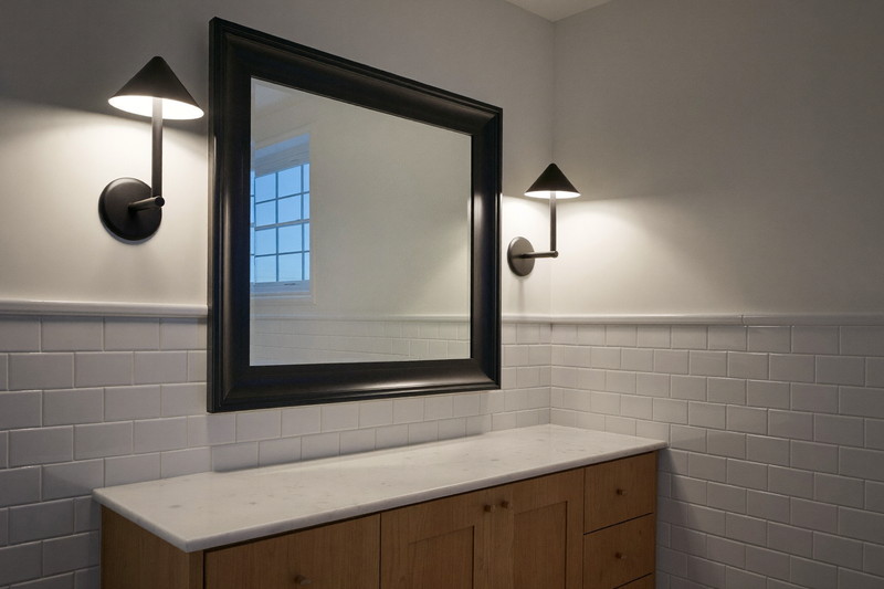 A calm finished small-town bathroom in soft evening light: matte black sconces glowing beside a rectangular black-framed mirror, pine vanity with a