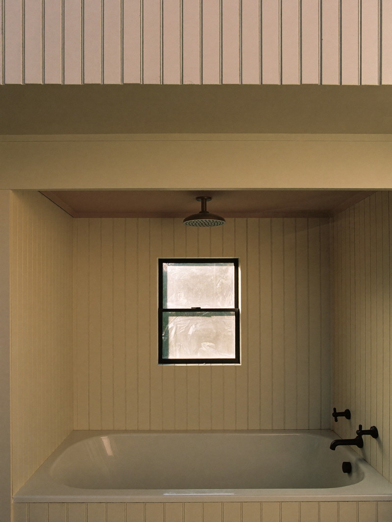 A renovated bathroom tucked under the roofline of a Millis cape: white tub, cream beadboard paneling, matte black fixtures, small window with frosted