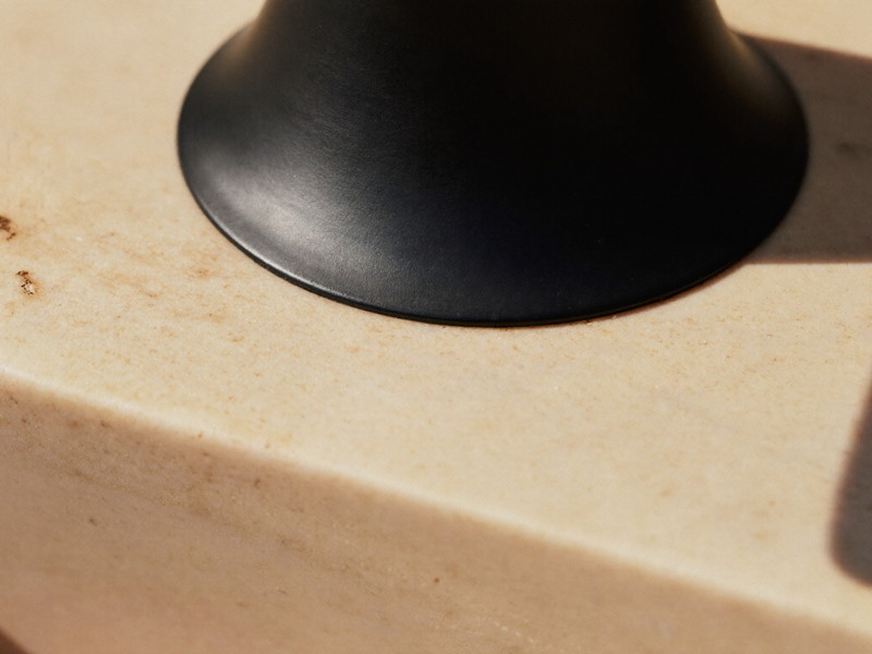 Macro photograph of creamy warm stone with faint speckles meeting a matte black smooth-faced faucet base, shallow depth of field, bright natural light
