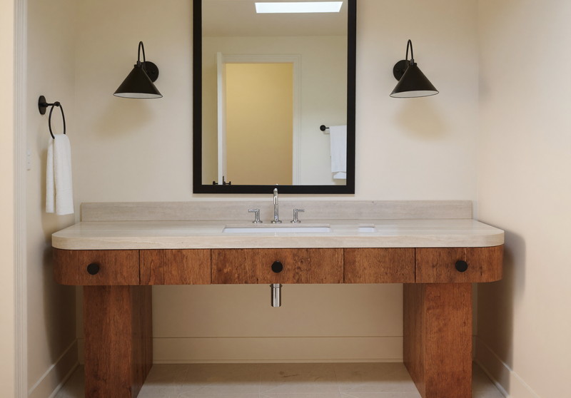 A single wide pine vanity with creamy stone top under one rectangular black-framed mirror, two matte black sconces, cream walls, tidy small-town bathroom
