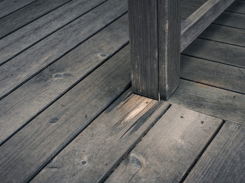 Close-up of an aged wooden deck corner on a well-kept small-town home before repair: weathered gray boards, one soft split board end, honest wear,