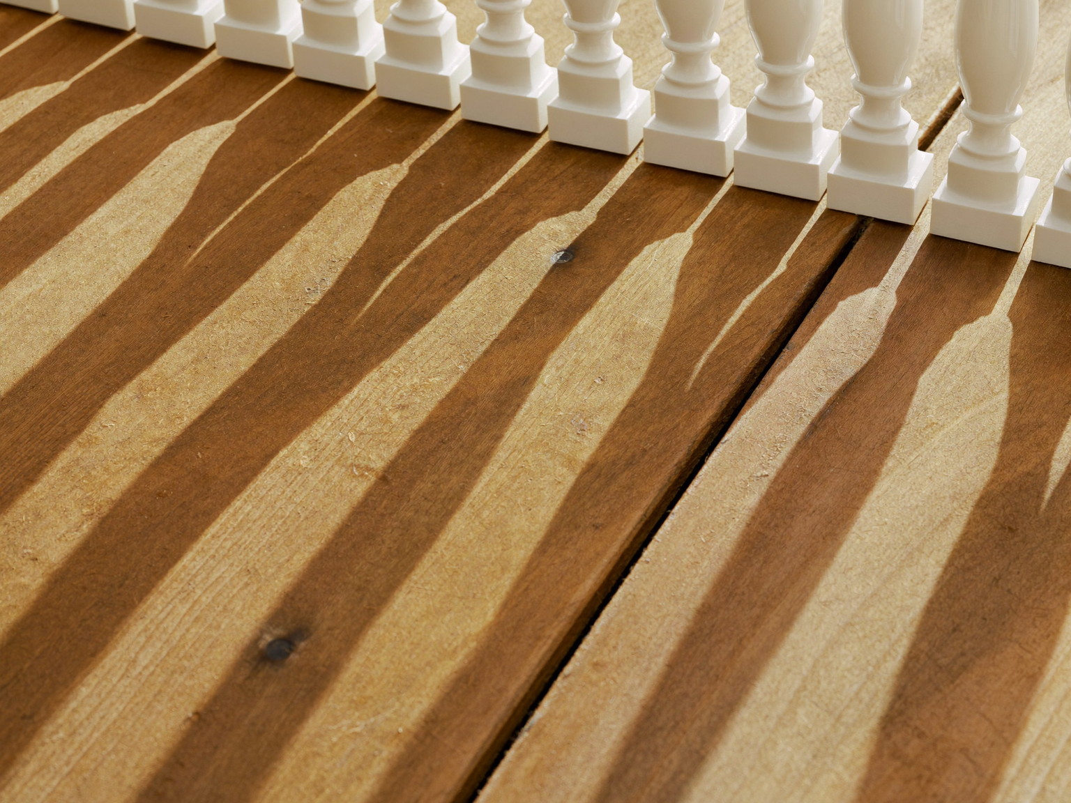 Freshly sealed pressure-treated deck boards in warm afternoon light: honey-toned surface with a soft sheen, white baluster shadows falling across the