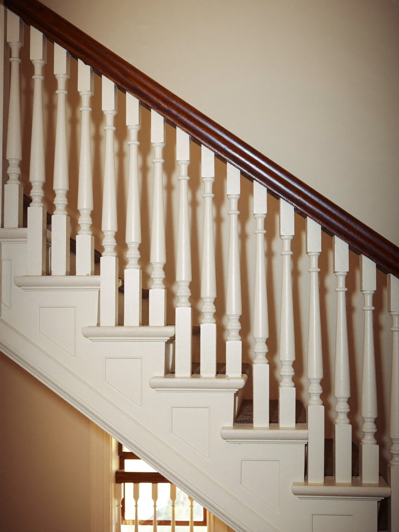 In a well-kept colonial stairwell, a repaired balustrade: one new crisp white baluster fitted among originals beneath a refinished dark pine handrail,