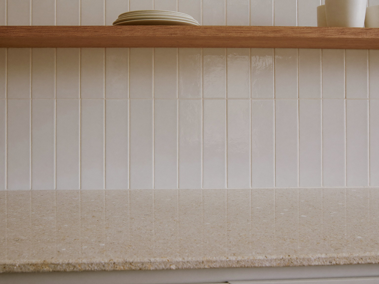 Macro photograph of a creamy speckled stone counter meeting a classic white subway tile backsplash, a slim pine shelf above holding plain white ceramics,