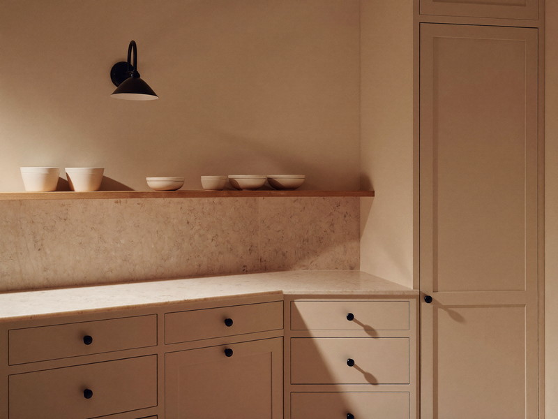 A finished small-town kitchen corner at evening: cream shaker cabinets with matte black knobs, creamy stone counter completely bare, pine shelf holding
