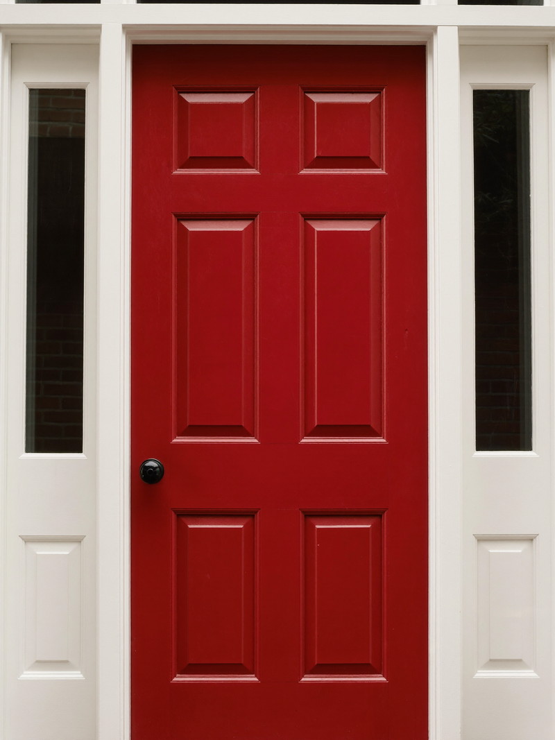A freshly painted barn-red front door on a well-kept Millis colonial: smooth satin finish, white paneled surround with clear glass sidelights, smooth