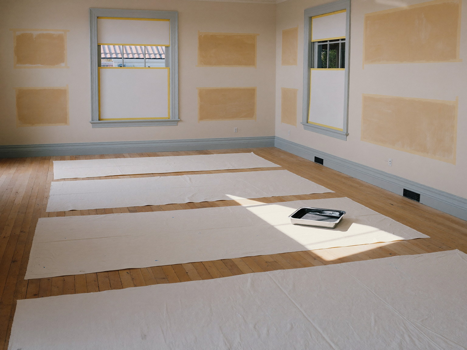Interior painting prep, seen from across a well-kept colonial room: clean canvas drop cloths spread over the wide pine floor, window casings masked with