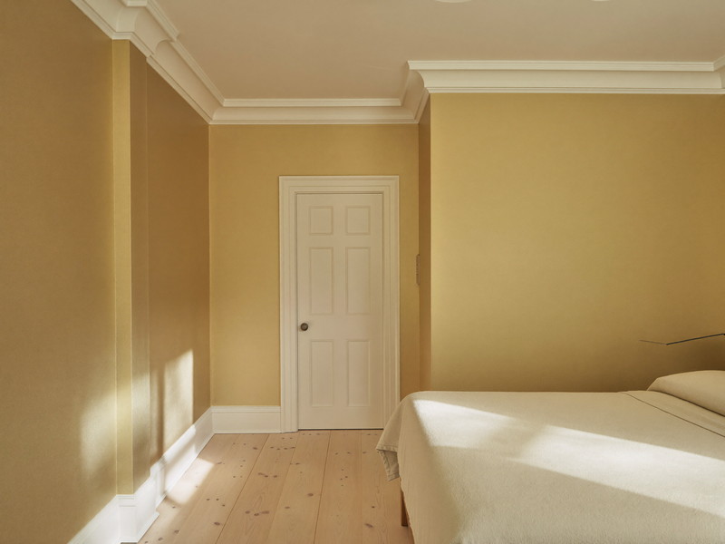 A repainted bedroom in a well-kept Millis colonial: soft wheat-gold walls, bright white trim and paneled door, smooth restored plaster in raking light,