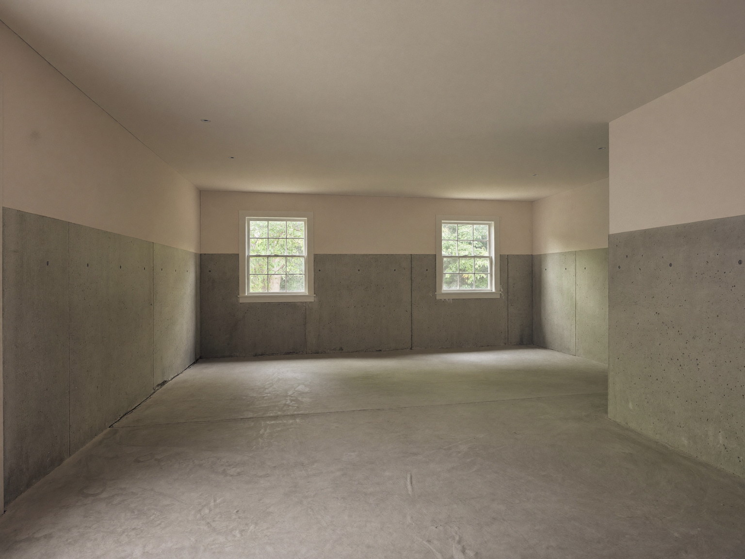 Before renovation: a clean, empty unfinished basement under an established suburban colonial with poured concrete foundation walls, smooth broom-swept