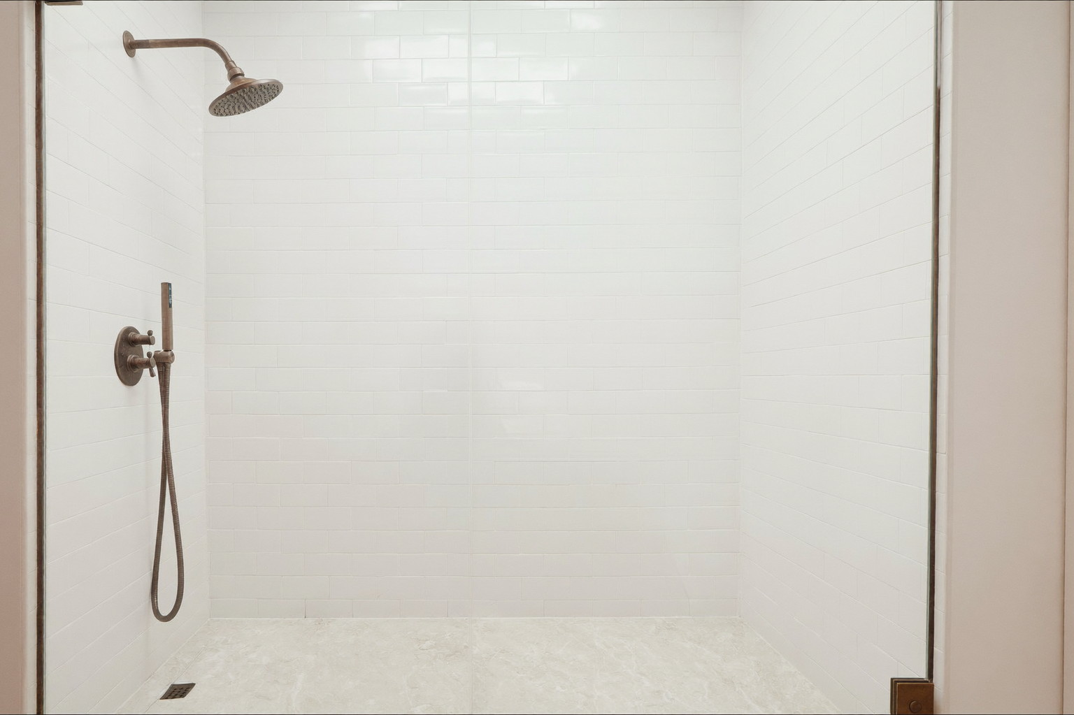 A wide view into a frameless-glass walk-in shower in a renovated Natick bathroom: warm white horizontal stacked tile, aged bronze rain head and controls,