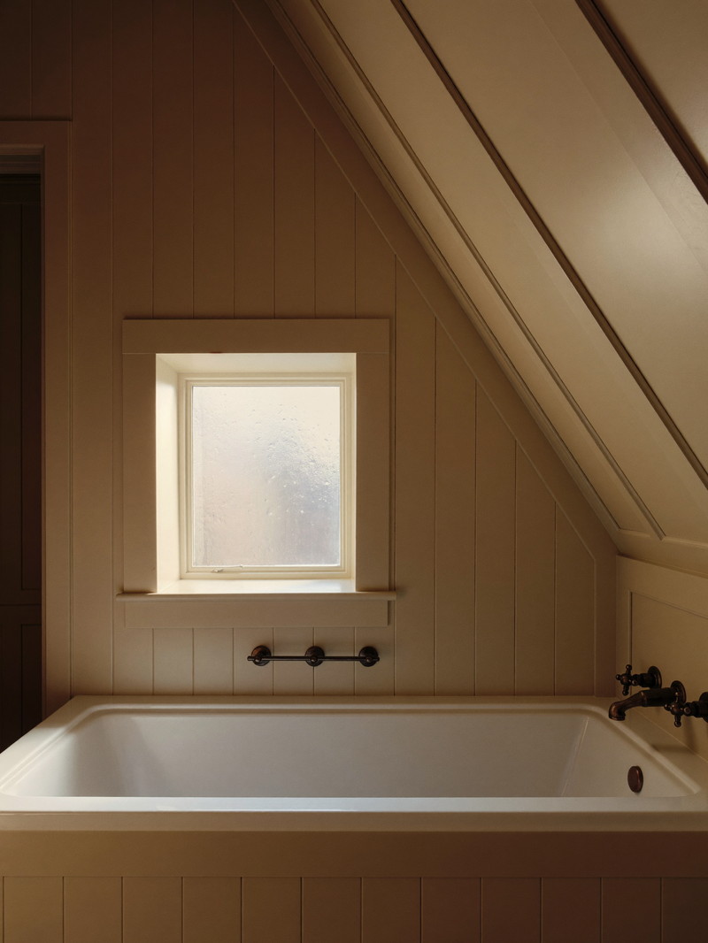 A renovated bathroom under a sloped eave in an established Natick cape: white tub, warm white paneling, aged bronze fixtures, small square window with