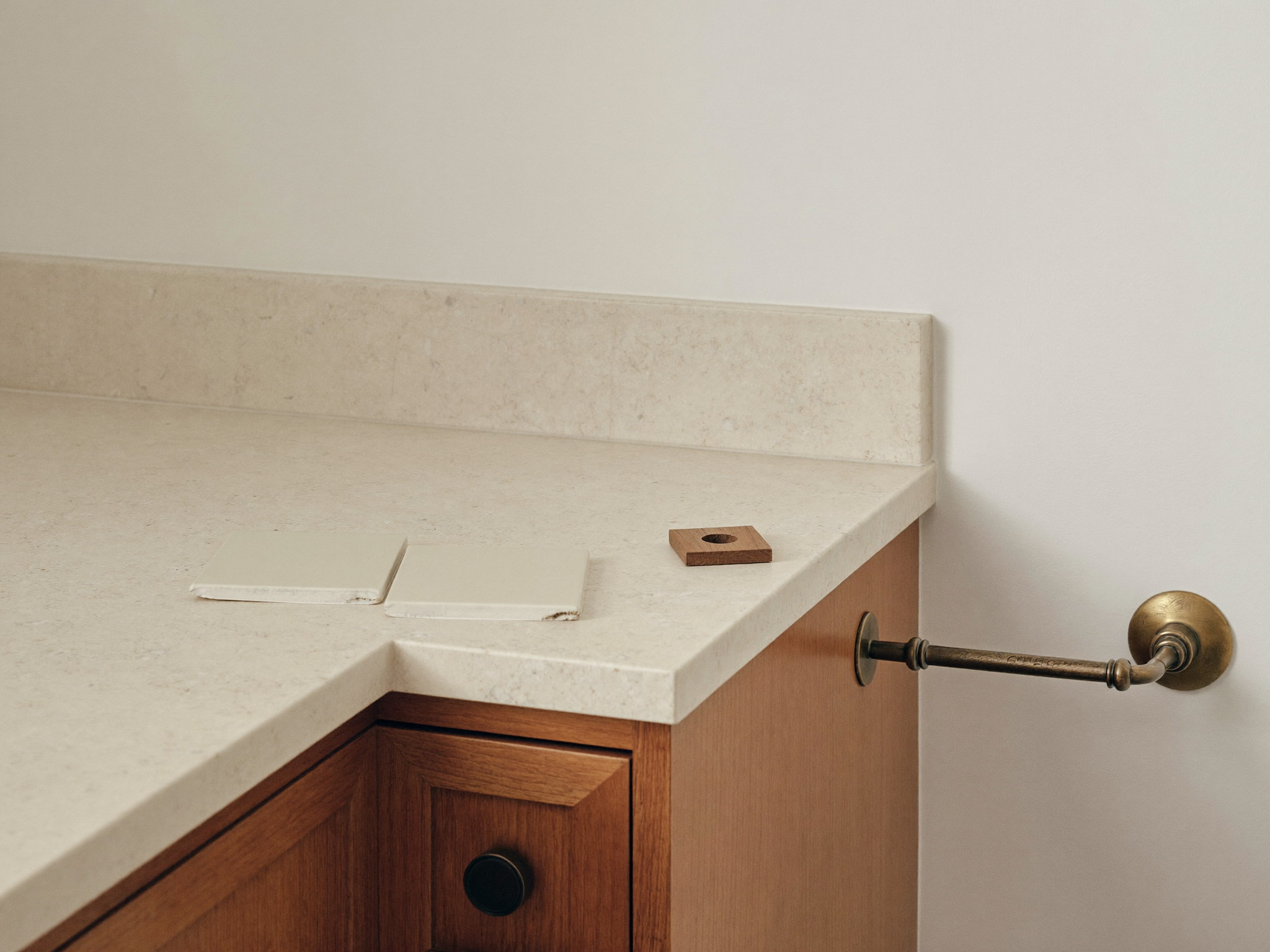 A corner of a cherry wood bathroom vanity in soft daylight: the pale quartzite top meeting a warm white wall, one loose warm white glazed tile and a small