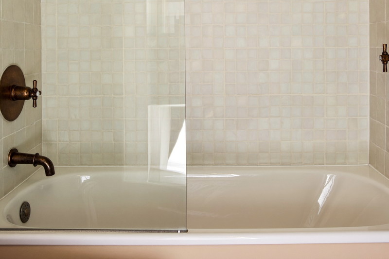 A warm tub and shower combination in a suburban family bathroom: white tub, warm white glazed tile surround, aged bronze fixtures, clear glass screen,